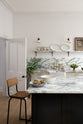 A quiet corner of a humble yet elegant off-white Classic English kitchen features a sink run with honed Arabescato marble worktop and splashback, a pantry blue kitchen island with a cooker, mini creamware wall lights, and a plumber’s stool. Adorned with artisan ceramics, a vintage painting, fresh greens, and grains, creating an intimate social and practical kitchen space.