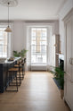 A humble yet elegant off-white Classic English kitchen features a Pantry blue kitchen island with a cooker, full height cabinets, and plumber’s stools. Adorned with artisan ceramics, a plate of fresh kiwis, a creamware pendant light, a grand tour desk lamp, and house plants, creating an intimate social and practical kitchen space.