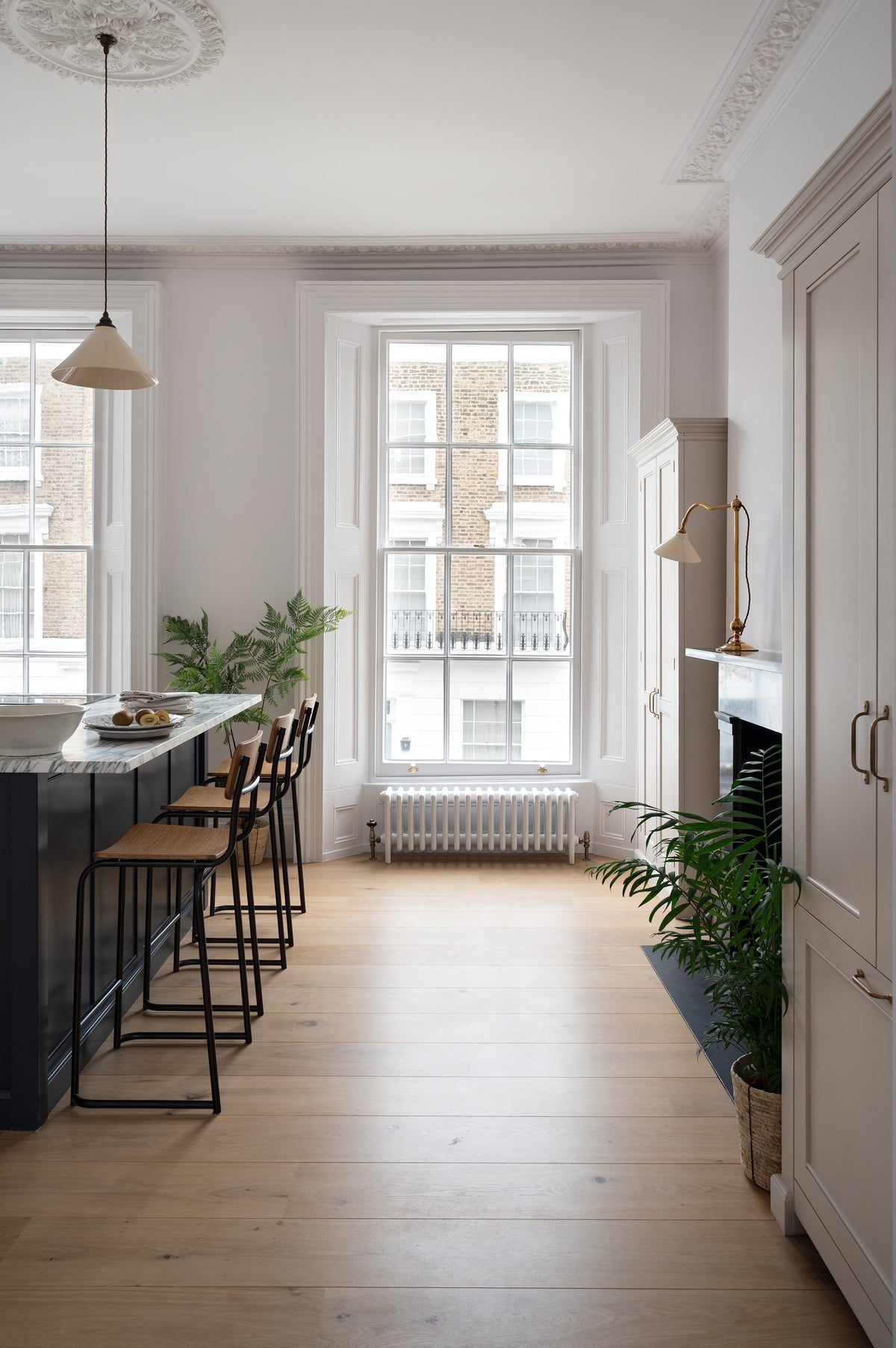 A humble yet elegant off-white Classic English kitchen features a Pantry blue kitchen island with a cooker, full height cabinets, and plumber’s stools. Adorned with artisan ceramics, a plate of fresh kiwis, a creamware pendant light, a grand tour desk lamp, and house plants, creating an intimate social and practical kitchen space.