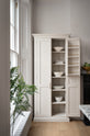 A humble yet elegant off-white Classic English kitchen features a full-height cabinet. Adorned with artisan ceramics, a bowl of fresh artichoke and house plants, creating an intimate social and practical kitchen space.