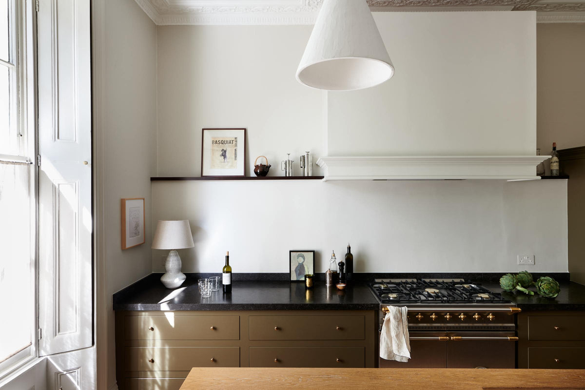 An elegant olive-brown classic kitchen features a cooker run topped with Belgian blue limestone, a kitchen island with a sink and a solid oak worktop, a shaker canopy, and a built-in shelf. The tall-ceilinged room is decorated with slender bottles of wine and vinegar, a pepper grinder, a vibrant green artichoke, and small, delicate framed illustrations.