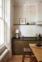 A quiet corner of an elegant olive-brown classic kitchen features a cooker run topped with Belgian blue limestone, a kitchen island with a sink and a solid oak worktop, a shaker canopy, and a built-in shelf. The tall-ceilinged room is decorated with slender bottles of wine and vinegar, a pepper grinder, a cocoa pod, and small, delicate framed illustrations.