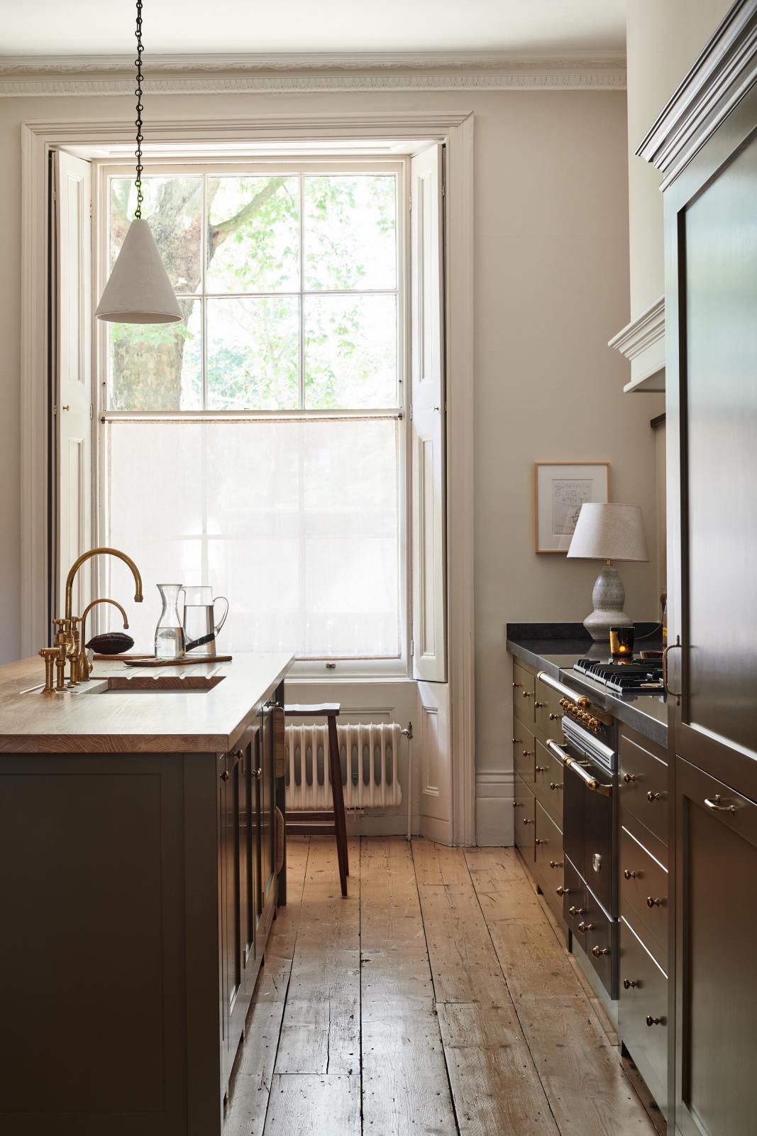 Looking at the wide windows in an elegant olive-brown classic kitchen in between the cooker run, topped with Belgian blue limestone and a kitchen island with a sink, a solid oak worktop, an aged brass Ionian tap, and wooden stools at the far edge.