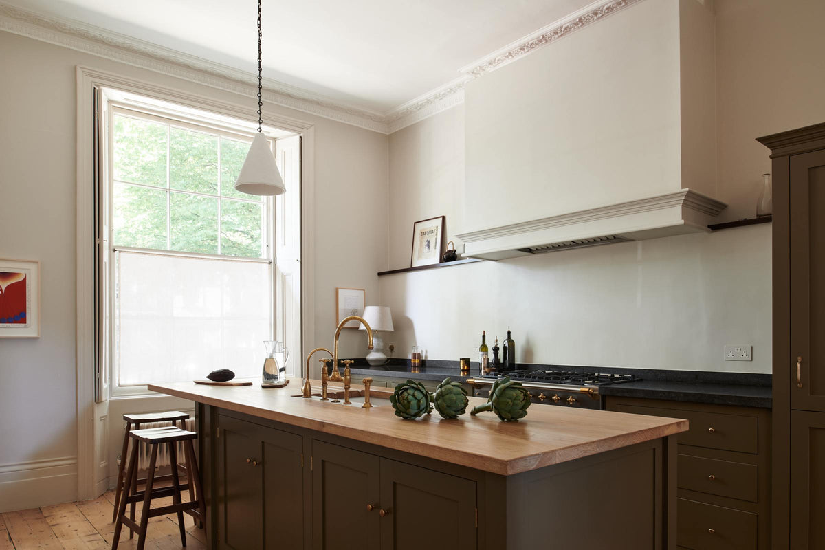 An oblique view of an elegant olive-brown classic kitchen featuring a cooker run topped with Belgian blue limestone, a shaker canopy, and a built-in shelf. At the centre is a kitchen island with a sink, a solid oak worktop, an aged brass Ionian tap, and a breakfast nook with wooden stools at the far edge. The tall-ceilinged room is decorated with slender bottles of wine and vinegar, a pepper grinder, a vibrant green artichoke, and small, delicate framed illustrations.