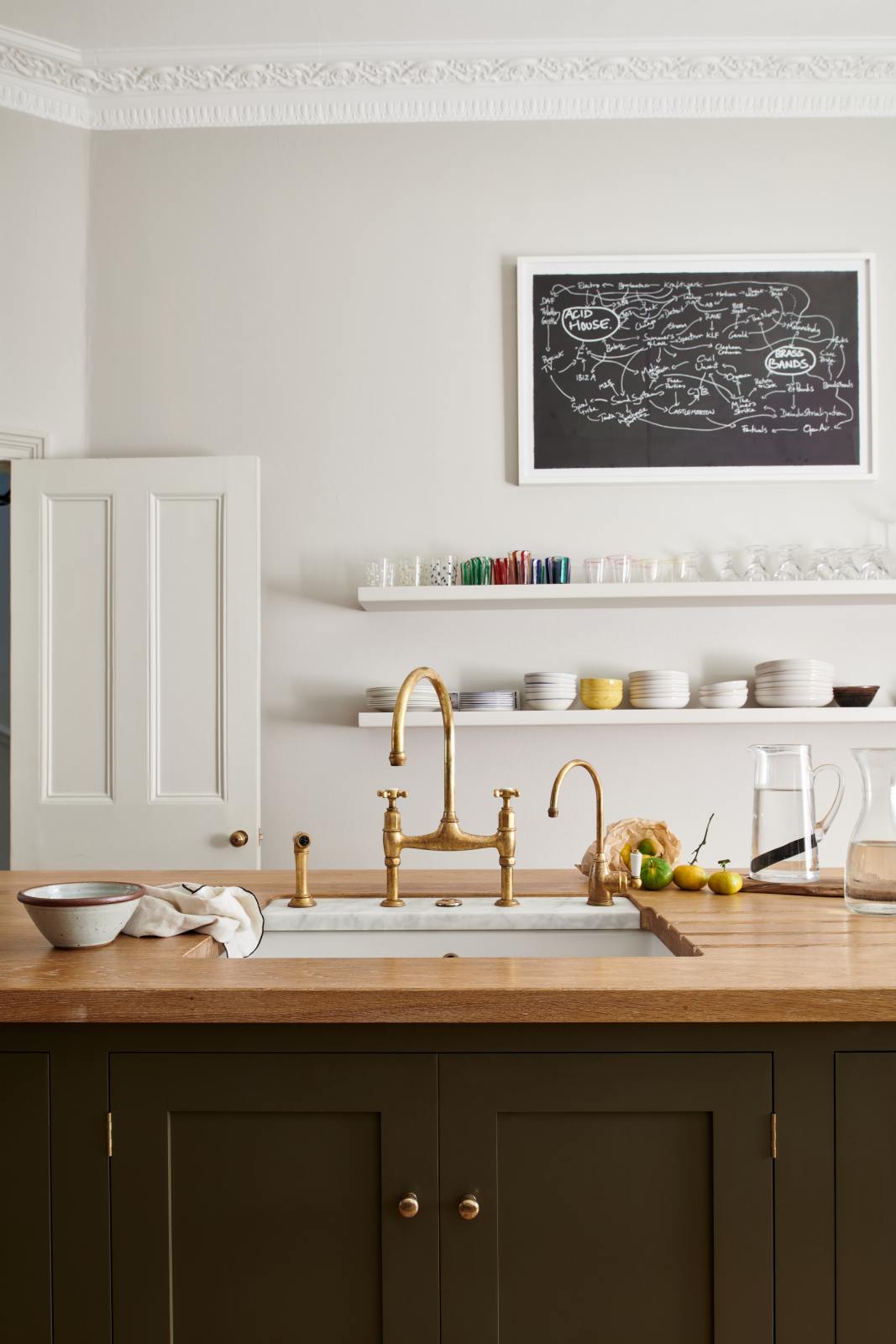 An elegant olive-brown classic kitchen features a kitchen island with a sink, a solid oak worktop, an aged brass Ionian tap, and a breakfast nook with wooden stools at the far edge. Long shelves line the wall, stacked with ceramic bowls, wine glasses, cocktail glasses, and drinking glasses. The tall-ceilinged room is decorated with slender water pitchers, glass milk, a bag of oranges, and big and colourful framed illustrations.