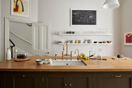 An elegant olive-brown classic kitchen features a kitchen island with a sink, a solid oak worktop, an aged brass Ionian tap, and a breakfast nook with wooden stools at the far edge. Long shelves line the wall, stacked with ceramic bowls, wine glasses, cocktail glasses, and drinking glasses. The tall-ceilinged room is decorated with slender water pitchers, glass milk, a bag of oranges, and big and colourful framed illustrations.