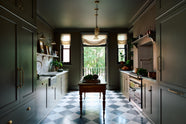 A grand and atmospheric olive green classic English kitchen features a marble worktop and splashback along the sink run, with a single marble sink and an aged brass Ionian tap. A cooker run with an overmantle is paired with an antique prep table on castors. Grand chequerboard marble tiles lead out onto a balcony overlooking green trees.