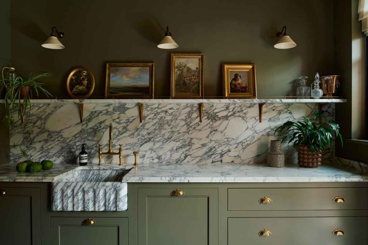 A grand and atmospheric olive green classic English kitchen features a marble worktop, splashback, and shelf along the sink run, a single fluted marble sink and an aged brass Ionian tap. Three beautiful Heirloom wall lights hang above gold-framed famous paintings, potted plants, and eccentric liquor bottles.
