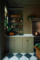 A grand and atmospheric olive green classic English kitchen features a cooker in the overmantle, an antique prep table on castors, and an open countertop cupboard filled with beautiful ceramic mugs, a teapot, potted plants, gold-foiled books, glasses, and framed artwork.