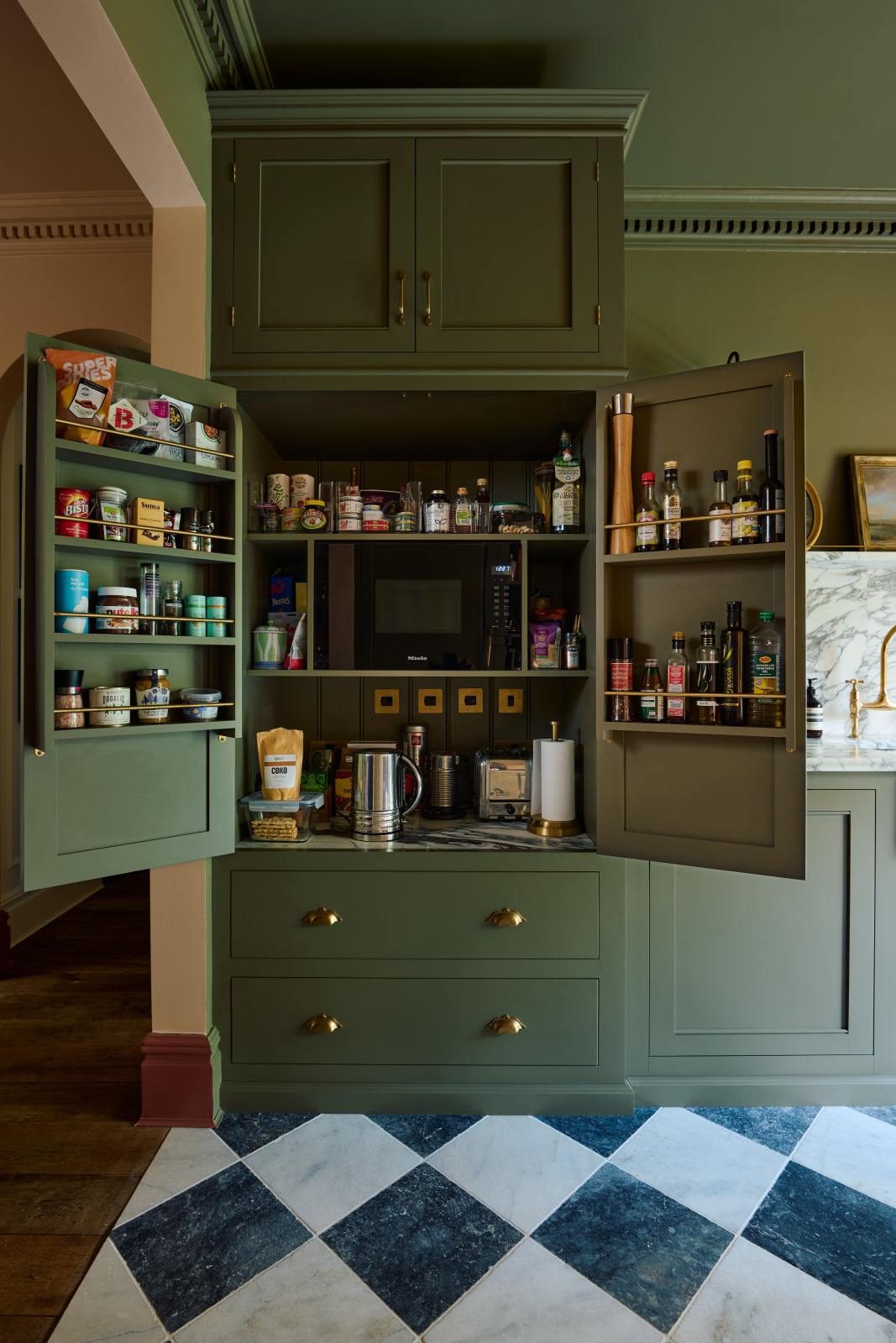 A grand and atmospheric olive green classic English kitchen features a marble worktop, splashback, and shelf along the sink run, a single fluted marble sink, and an aged brass Ionian tap. An open full-height pantry cabinet takes centre stage, with assorted shelves and open cabinets that store a variety of cooking essentials.