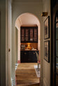Across a tall-ceiling hallway and through the archway is a small, printer’s black classic English café section with glazed, reeded cupboards and cabinets with aged copper worktop and splashback. An elegant and sophisticated area in the dining room, complete with a silver coffee machine with a milk frother, as well as wine, cocktail, and scotch glasses.