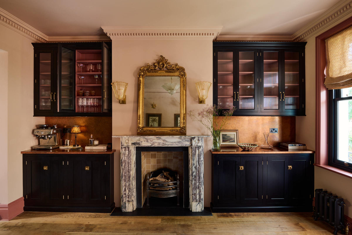 A grand dining room with a printer’s black, classic English café-style nook, reeded glazed cupboards, and aged copper worktops and splashback. A gilded mirror rests above a marble-framed fireplace, framed by amber frilled sconces.