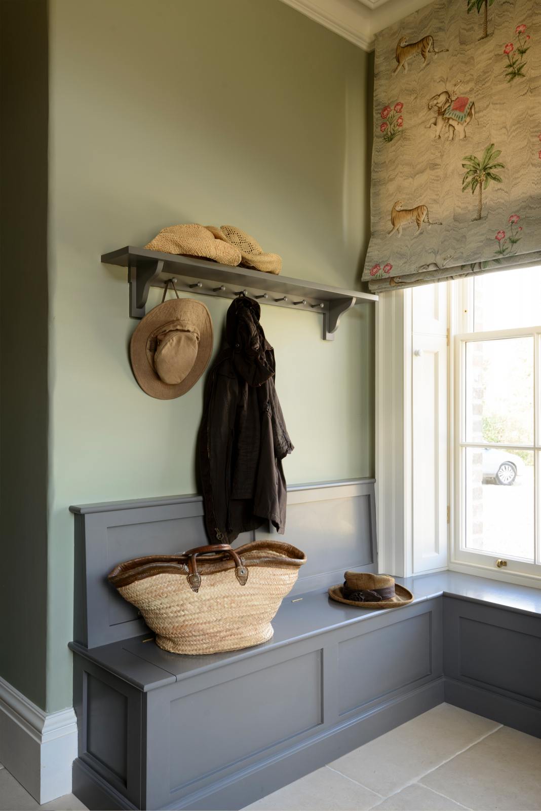 A quiet corner of a functional, muted-grey boot room features an L-shaped bench and a shelf with a peg rail. Beautifully decorated with thick coats, a stack of hats, and a woven shoulder bag, this creates a practical and family-oriented space.