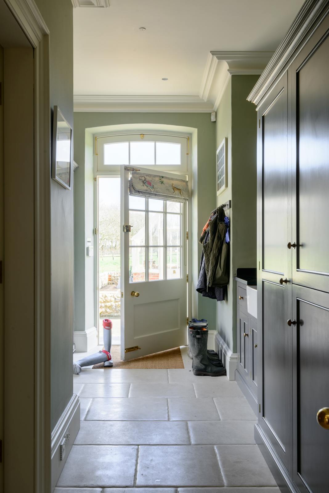A functional muted-grey boot room features an L-shaped bench, shelves, a peg rail, a small sink run with a single Belfast Ionian sink, a black granite worktop, and full-height storage cupboards. Beautifully decorated with thick coats and muddy boots, this space leads into the back garden, creating a practical and family-oriented area.