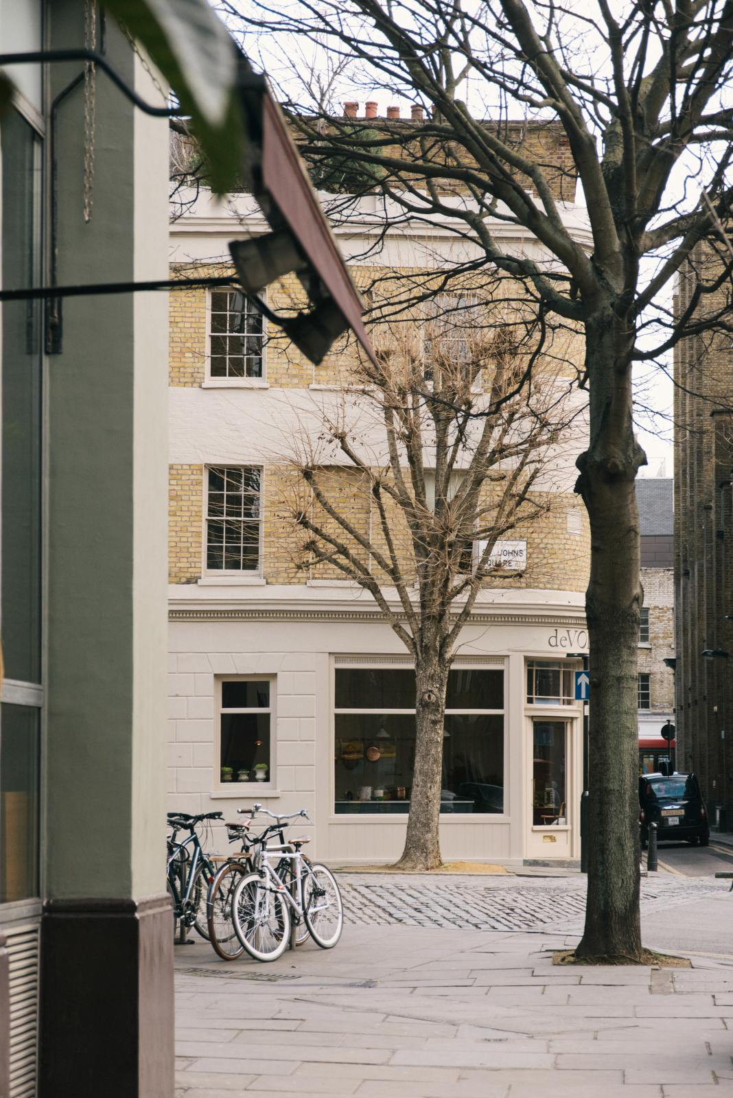 Beyond winter trees, cobblestones, and bike racks is a tall historical townhouse located at a corner in the heart of London, which has been beautifully renovated in a shade known as Princelet pink. The building still showcases its original brick texture. Above the entrance door is the deVOL logo in black with a gold-rimmed shadow.