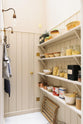 A narrow, high-ceilinged pantry painted beige, featuring four shelves filled with dried ingredients, copper pots and pans, jars of jam, a large can of tomato paste, and an aged brass hanging rail.