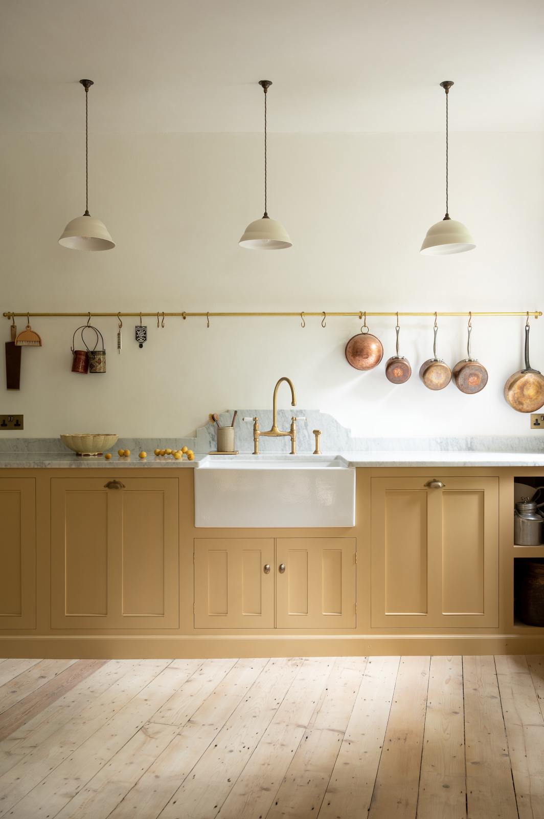 This charming blush yellow, classic English kitchen features a Carrara marble countertop, an ornate backsplash, and an aged brass tap. Decorated with copper pots, vintage ceramics, and small wooden cleaning supplies, it creates an elegant and tidy atmosphere.
