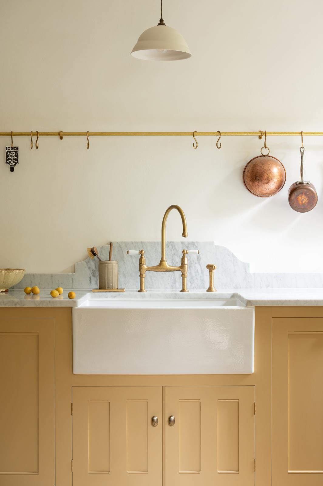 A closer look at a charming blush yellow, classic English kitchen features a Carrara marble countertop, an ornate backsplash, and an aged brass tap. Decorated with copper pots, vintage ceramics, and small wooden cleaning supplies, it creates an elegant and tidy atmosphere.