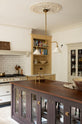 An oblique view of a lovely and simple blush yellow, classic English kitchen features a cooker run with Carrara marble worktop, two open countertop cupboards, a white gas stove inside a chimney breast, porcelain splashback, a purple-brown kitchen island with a solid oak worktop and aged brass Mayan taps. A deVOL gaselier light and a collection of muted ceramics and tall wildflowers create an elegant and inviting atmosphere in the room.