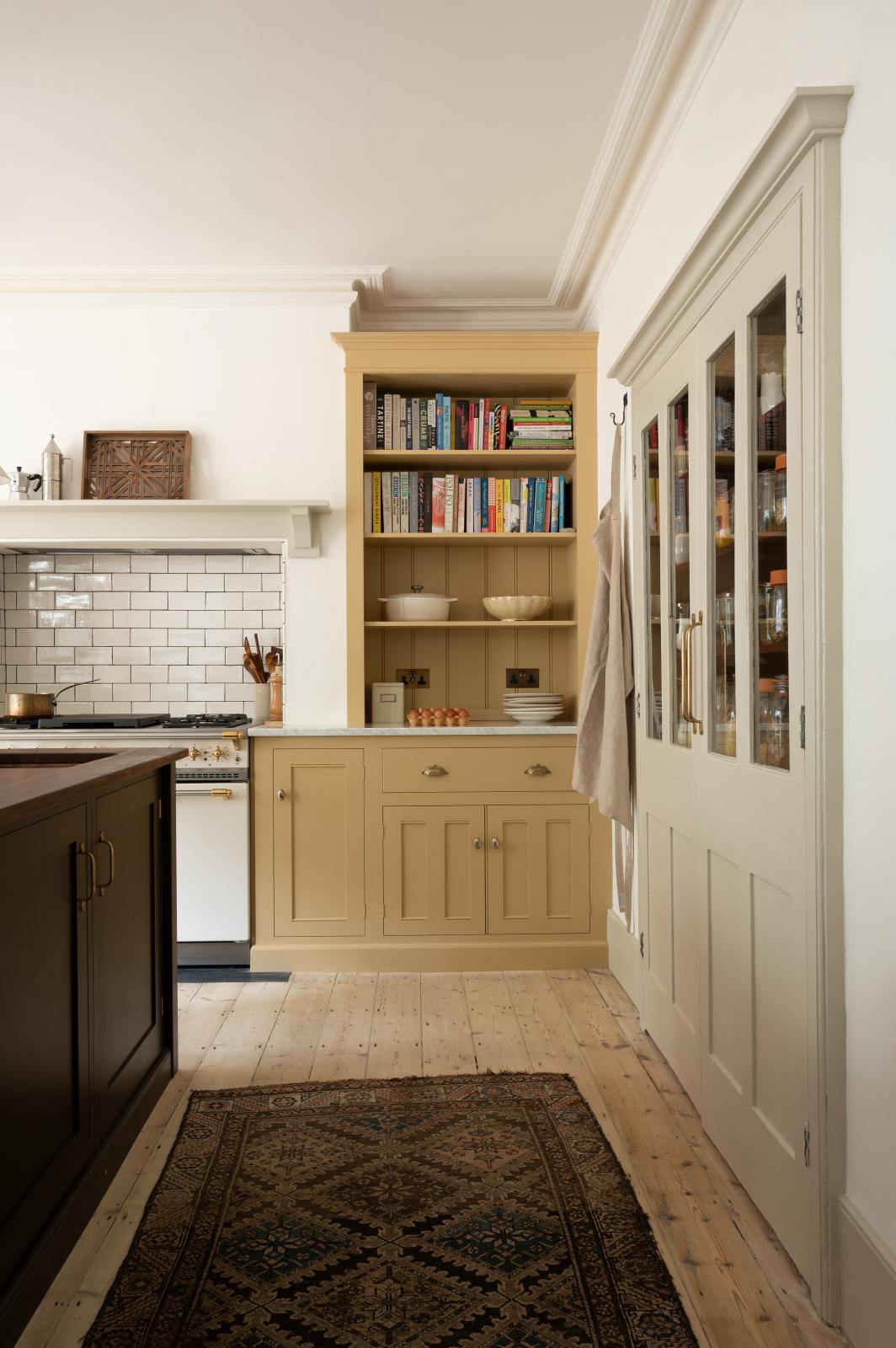 Nestled between a purple-brown kitchen island with a solid oak worktop and a shallow pantry cupboard is a classic English kitchen cooker in blush yellow. A collection of books, muted ceramics, and a carton of eggs enhance the inviting atmosphere.