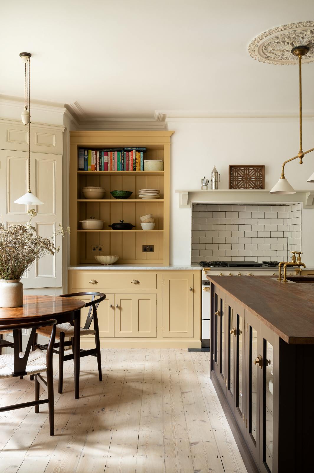 A lovely and simple blush yellow, classic English kitchen features a cooker run with Carrara marble worktop, two open countertop cupboards, a white gas stove inside a chimney breast, porcelain splashback, a purple-brown kitchen island with a solid oak worktop and aged brass Mayan taps. A circular coffee table with matching chairs, a deVOL gaselier light, and a collection of muted ceramics and tall wildflowers create an elegant and inviting atmosphere in the room.