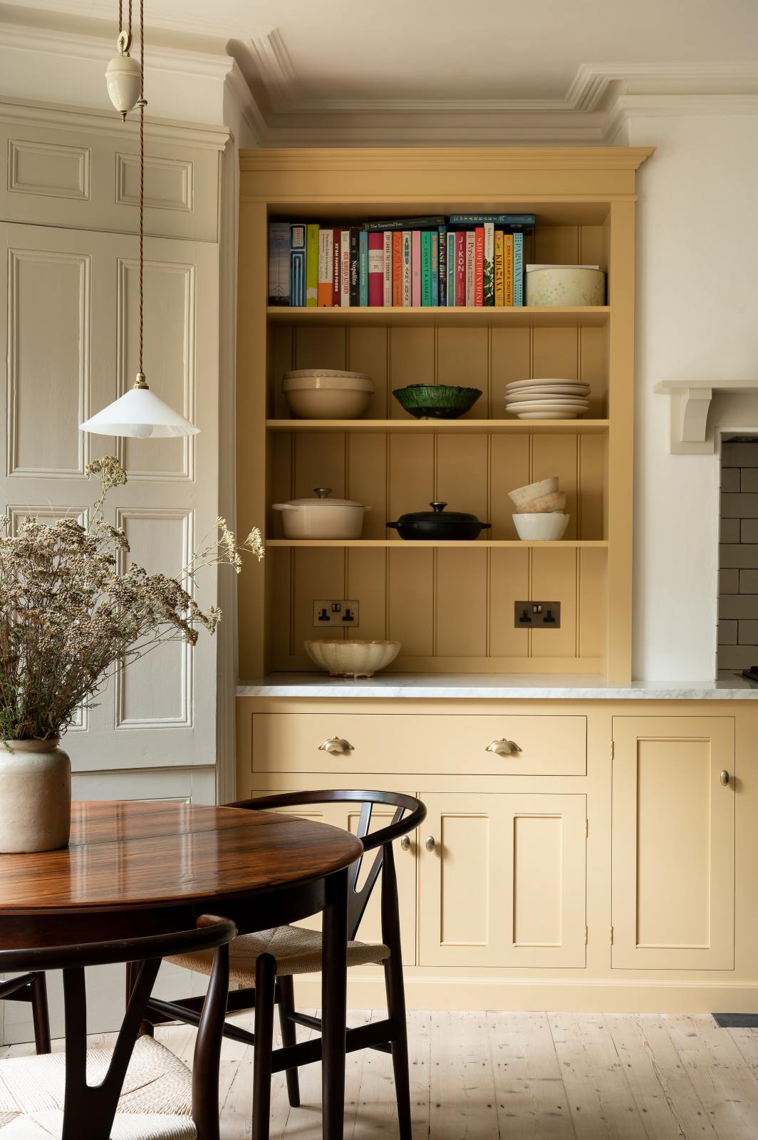 A lovely and simple blush yellow, classic English kitchen features a cooker run with Carrara marble worktop, two open countertop cupboards, a white gas stove inside a chimney breast, porcelain splashback, a purple-brown kitchen island with a solid oak worktop and aged brass Mayan taps. A circular coffee table with matching chairs, and a collection of muted ceramics and tall wildflowers create an elegant and inviting atmosphere in the room.