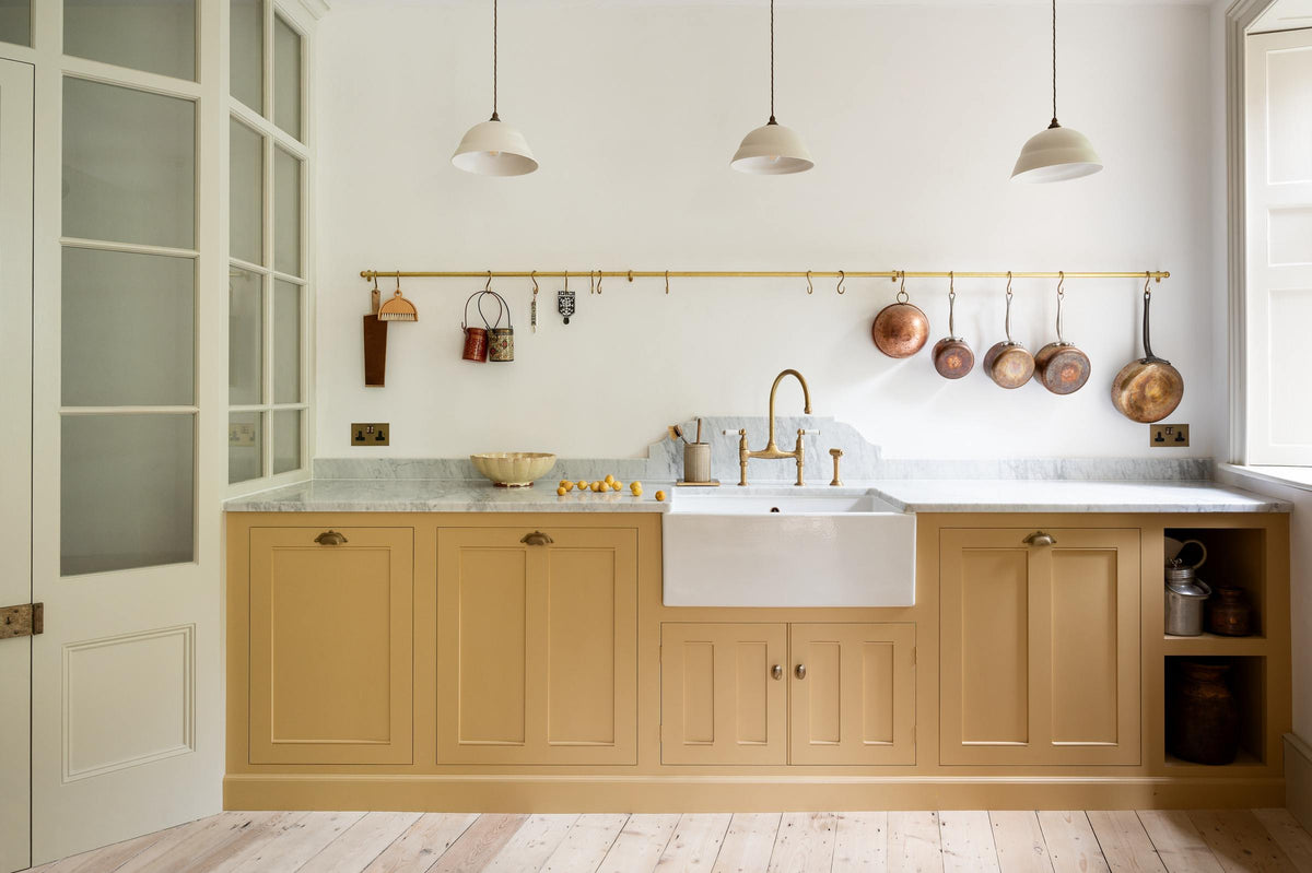 A charming blush yellow, classic English kitchen features a Carrara marble countertop, an ornate backsplash, and an aged brass tap. Decorated with copper pots, vintage ceramics, and small wooden cleaning supplies, it creates an elegant and tidy atmosphere.