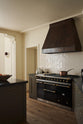 A spacious olive-green classic kitchen features a cooker run with a large black gas stove, a kitchen island with a Belgian blue limestone worktop, a huge rustic metal canopy and white splashback tiles.