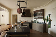 A spacious olive-green classic kitchen features a large island with a Belgian blue limestone worktop, a single sink, and an antique silver Ionian tap. Above, a deVOL pot hanger takes centre stage. The classic cooker run showcases a large black gas stove, a substantial canopy, white splashback tiles, and an heirloom plate rack displaying colour-coordinated muted greens and dark blue ceramic plates.