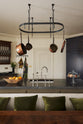 A spacious and sophisticated classic kitchen in olive green features a large kitchen island topped with Belgian blue limestone, a single sink, and an antique silver Ionian tap. In front of the kitchen island is a large vintage dining table with a bench decorated with green pillows. Above the island, a deVOL pot hanger takes centre stage.