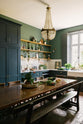 The spacious kitchen in the renovated rectory features a blue L-shaped countertop, a mix of cabinetry against green walls, and a metro tile backsplash. At the centre, a copper-topped table flanked by a vintage bench and chairs is complemented by an elegant antique chandelier.