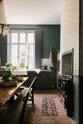 The spacious kitchen in the renovated rectory features a blue L-shaped countertop, a mix of cabinetry against green walls, and a metro tile backsplash. At the centre, a copper-topped table flanked by a vintage bench and chairs is complemented by an elegant antique chandelier.