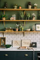 The deep blue base cupboards contain an under-counter oven and are topped with Carrara marble. A lovely white metro tile splashback extends up the wall to meet the first three wooden shelves, each supported by beautiful handmade aged brass brackets. The walls are painted a muted dark green, and the shelves display an array of trinkets, plants, and artwork.