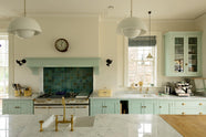 A humble and calming turquoise, classic English kitchen featuring a cooker and sink run with a Carrara marble worktop, a suspended false chimney breast, a glazed wall cupboard, and a kitchen island with Carrara marble on one side and solid oak on the other. Adorned with symmetrical hanging lights, butter-yellow ceramics, containers, and aged brass Ionian and Mayan taps, it creates a warm and simple home kitchen.