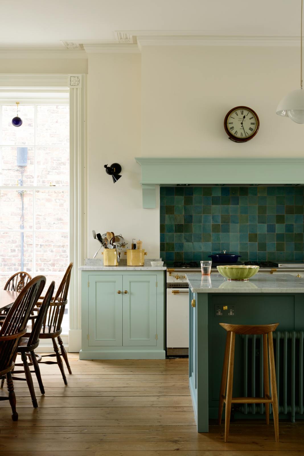 A corner of a humble and calming turquoise, classic English kitchen featuring a cooker and sink run with a Carrara marble worktop, a suspended false chimney breast, a glazed wall cupboard, and a kitchen island with Carrara marble on one side and solid oak on the other. Adorned with symmetrical hanging lights, butter-yellow ceramics, deVOL bum stools and ocean porcelain tiles, it creates a warm and simple home kitchen.