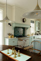 An oblique view of a humble and calming turquoise, classic English kitchen featuring a cooker and sink run with a Carrara marble worktop, a suspended false chimney breast, a glazed wall cupboard, and a kitchen island with Carrara marble on one side and solid oak on the other. Adorned with symmetrical hanging lights, butter-yellow ceramics, golden candle stick holders and ocean porcelain tiles, it creates a warm and simple home kitchen.