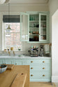 A corner of a humble and calming turquoise, classic English kitchen featuring a cooker and sink run with a Carrara marble worktop, a suspended false chimney breast, a glazed wall cupboard, and a kitchen island with a solid oak worktop. Adorned with symmetrical hanging lights, artisan ceramics, silver coffee machines and fresh eggs, it creates a warm and simple home kitchen.