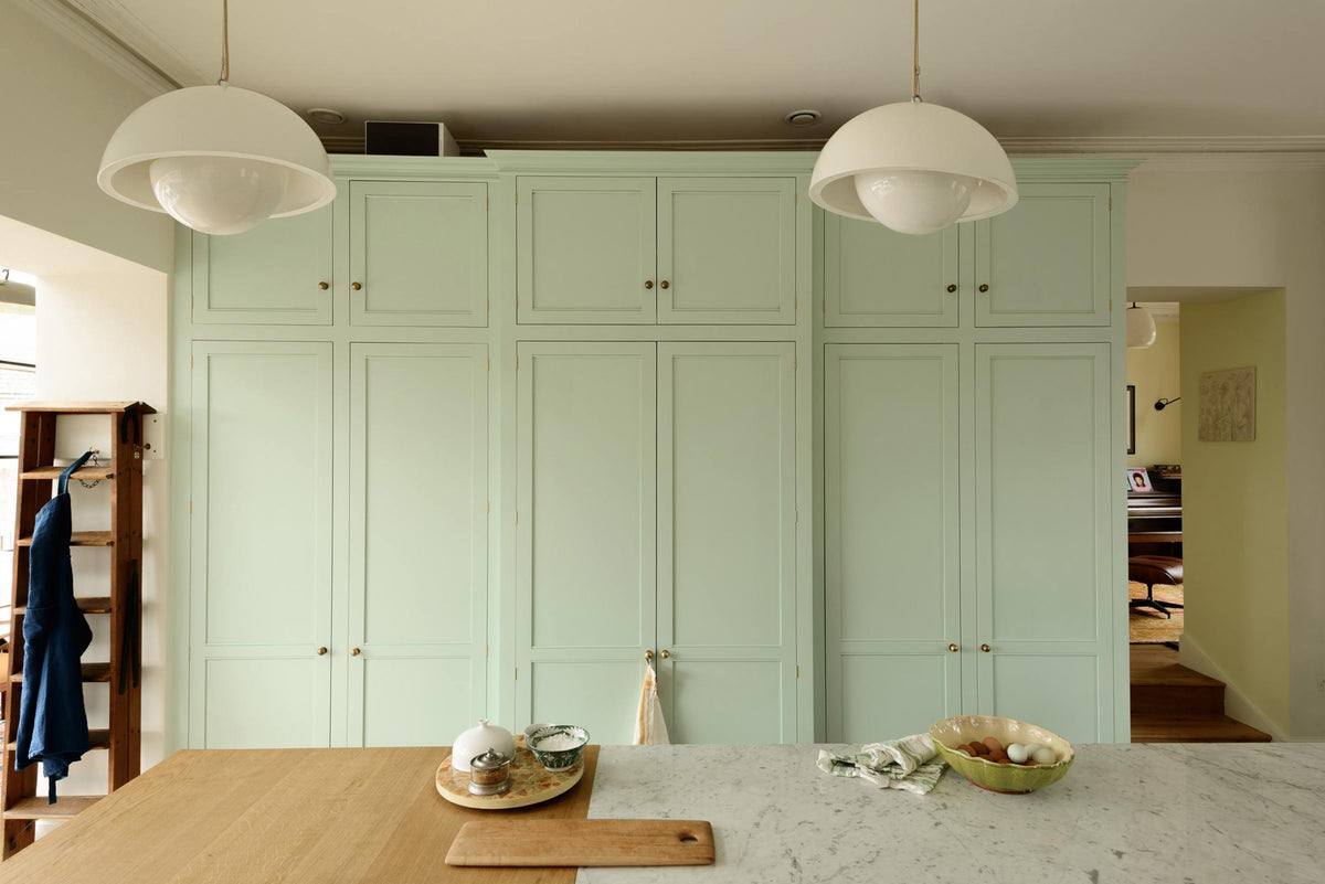 A humble and calming turquoise, classic English kitchen featuring a large full-height pantry cupboards with top cabinets, a wooden ladder, and a kitchen island with Carrara marble on one side and solid oak on the other. Adorned with symmetrical hanging lights, artisan ceramics, a wooden chopping board, and a large bowl of fresh eggs, it creates a warm and simple home kitchen.