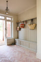 An oblique view of a modest foyer featuring a wooden compartment bench, hooks, and a shelf painted in a muted sage green, complemented by the pale terracotta parquet floor, wicker bags, stray hats, and green and white striped slippers, adds to the coastal aesthetic.