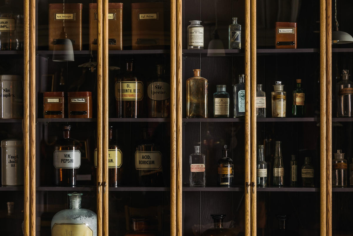 A closer look at a cool and elegant Haberdasher’s collection featuring a pantry run with glazed countertop cabinets. Adorned with vintage items, assorted containers, and bottles, this space creates an atmospheric social bar.