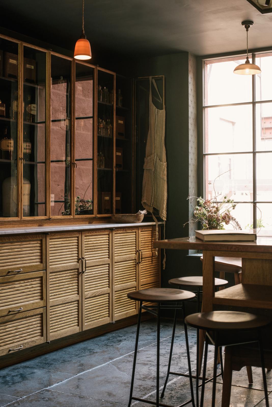 An oblique view of a cool and elegant Haberdasher’s collection features a large prep table with a copper worktop, a pantry run with tambour base cabinets, and glazed countertop cabinets. Adorned with vintage furniture and items, artisan ceramics, the deVOL porcelain light collection, Creedy stools, assorted containers, bottles, and a stunning bouquet of dried flowers, this space creates an atmospheric social bar.