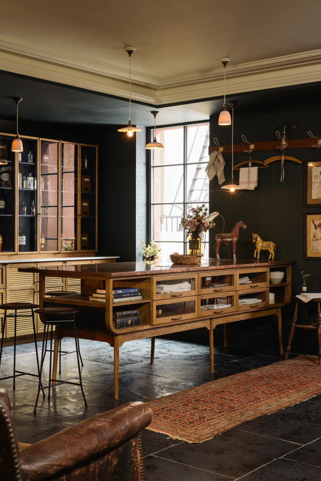 An oblique view of a cool and elegant Haberdasher’s collection features a large prep table with a copper worktop, a pantry run with tambour base cabinets, glazed countertop cabinets, and a freestanding pantry with glazed doors. Adorned with vintage furniture and items, artisan ceramics, the deVOL porcelain light collection, Creedy stools, assorted containers, bottles, and a stunning bouquet of dried flowers, this space creates an atmospheric social bar.