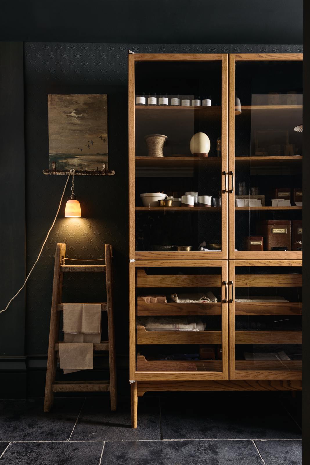 A quiet corner of a cool and elegant Haberdasher’s collection features a freestanding pantry with glazed doors. Adorned with vintage furniture and items, artisan ceramics, a plug-in porcelain beaker lights, assorted containers, and candles, this space creates an atmospheric social bar.