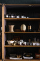 A closer look at a cool and elegant Haberdasher’s collection featuring a freestanding pantry with glazed doors. Adorned with vintage items, artisan ceramics, assorted containers, and candles, this space creates an atmospheric social bar.