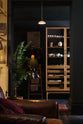 A quiet corner of a cool and elegant Haberdasher’s collection features a freestanding pantry with glazed doors. Adorned with vintage furniture and items, artisan ceramics, a plug-in porcelain beaker lights, assorted containers, and candles, this space creates an atmospheric social bar.