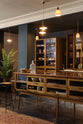 An oblique view of a cool and elegant Haberdasher’s collection features a large prep table with a copper worktop, a pantry run with tambour base cabinets, glazed countertop cabinets, and a freestanding pantry with glazed doors. Adorned with vintage furniture and items, artisan ceramics, the deVOL porcelain light collection, Creedy stools, assorted containers, and bottles, this space creates an atmospheric social bar.
