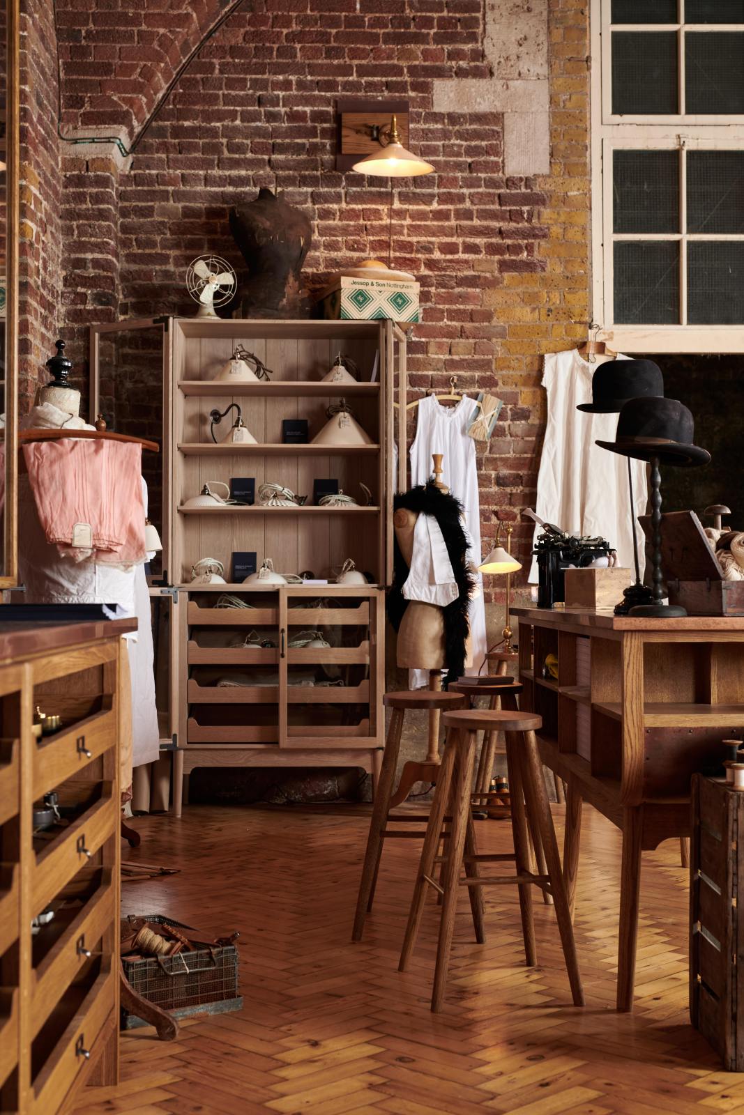 A handsome range of oak furniture and lighting from the deVOL Haberdasher’s collection features a kitchen island, countertop cupboards, and a glazed freestanding pantry. Adorned with deVOL Heirloom accessories, Helen stools, vintage furniture and items, dressmaker mannequins, and 1900s tailor tools, this space creates a beautiful homage to the English gentlemen’s haberdashery inspiration in a bricked wall showroom.