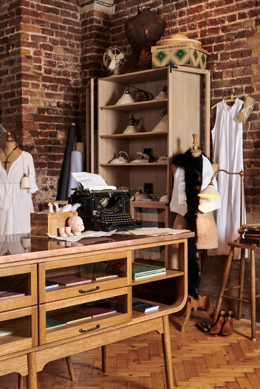 A close and oblique look at a handsome range of oak furniture and lighting from the deVOL Haberdasher’s collection features a kitchen island, and a glazed freestanding pantry. Adorned with deVOL Heirloom accessories, Helen stools, vintage furniture and items, dressmaker mannequins, and 1900s tailor tools, this space creates a beautiful homage to the English gentlemen’s haberdashery inspiration in a bricked wall showroom.