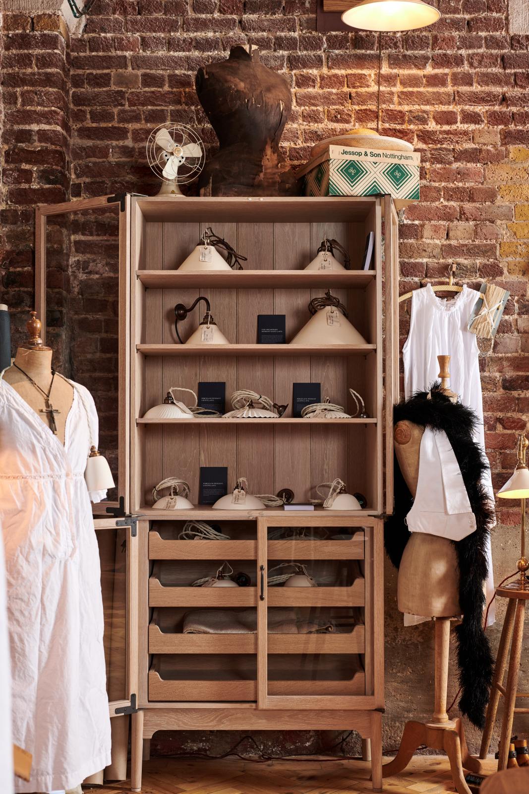 A closer look at the glazed freestanding pantry cupboard from the deVOL Haberdasher’s collection reveals a kitchen island and countertop cupboards. Adorned with deVOL Heirloom accessories, creamware pendant lights, porcelain pendant lights, Helen stools, vintage furniture and items, dressmaker mannequins, and 1900s tailor tools, this space creates a beautiful homage to the English gentlemen’s haberdashery inspiration in a bricked wall showroom.