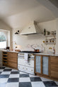 An oblique view of a grand and quaint Haberdasher’s kitchen features a sink and cooker run with a Paonazzo marble worktop and decorative vanity cut. Beautifully decorated with a white gas cooker with golden accents, a matching canopy, a deVOL grand tour desk lamp, vintage furniture, beloved chapel chairs, a skylight, and a cloth lampshade, this kitchen creates a humble and functional space with a touch of elegance.
